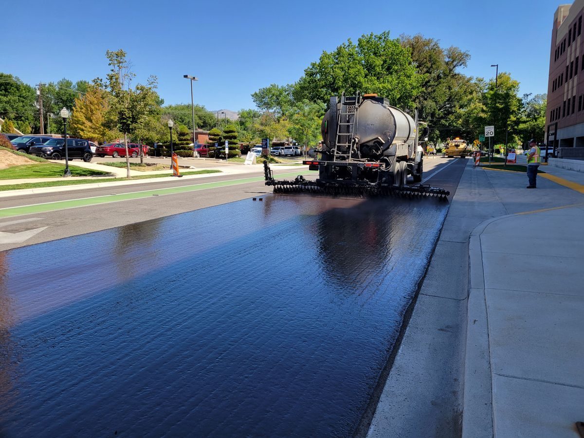 Road treatment truck working on a street beside marked parking areas.