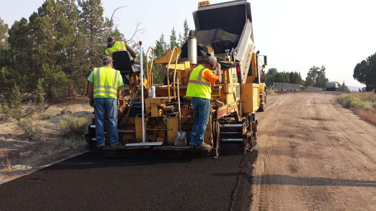 Road workers in reflective vests operating paving equipment.