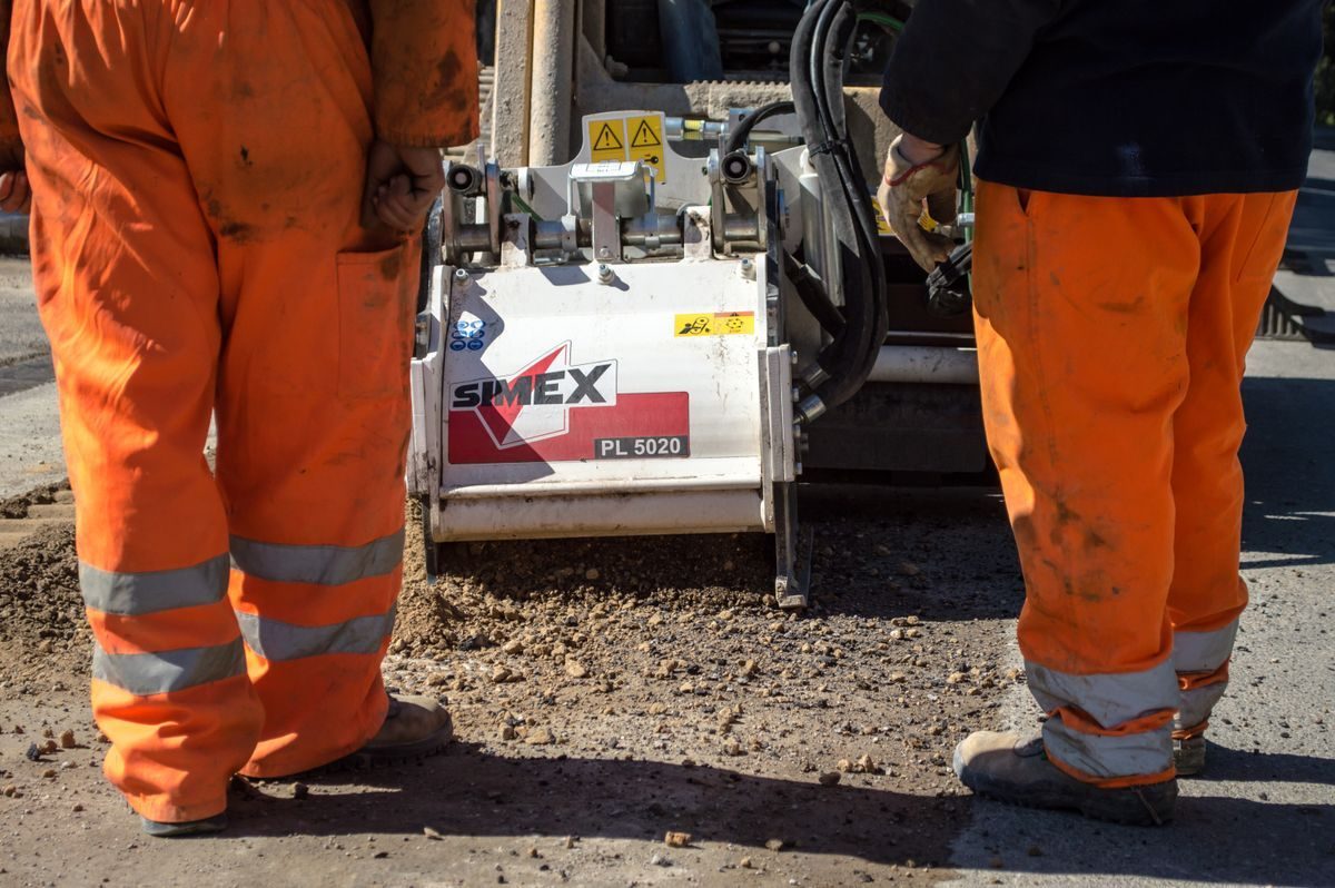 Workers in orange safety trousers beside compact roadwork equipment.