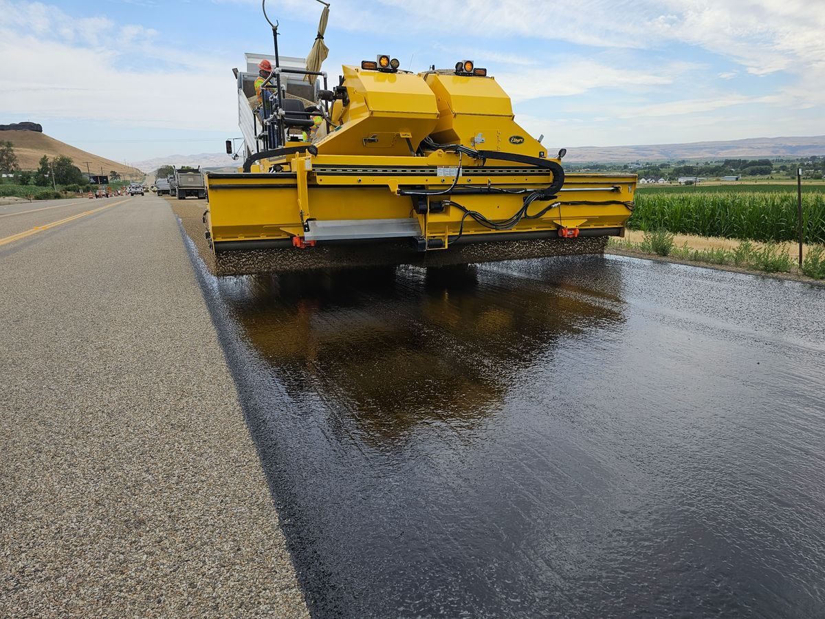 Yellow road machine applying a dark surface treatment along a rural roadway.