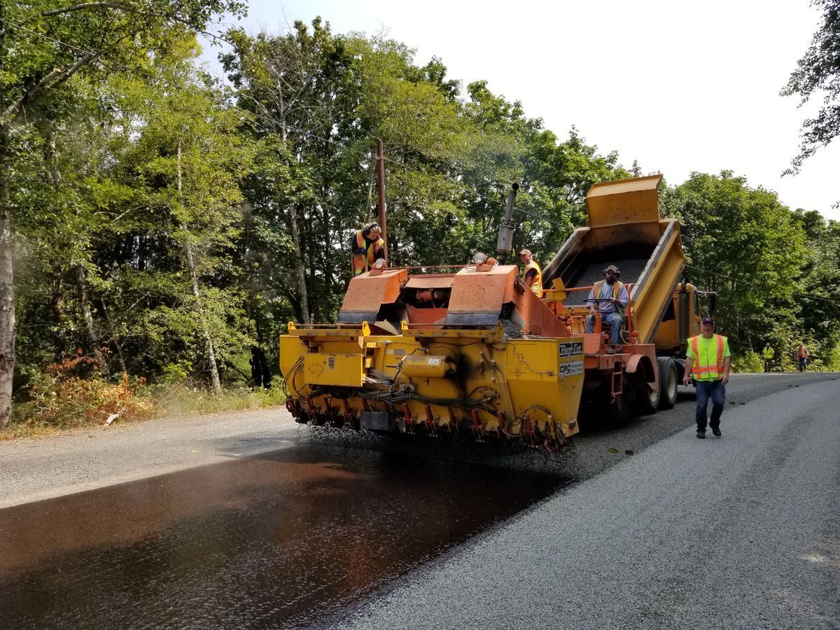 Road crew working beside paving equipment on a tree-lined road.