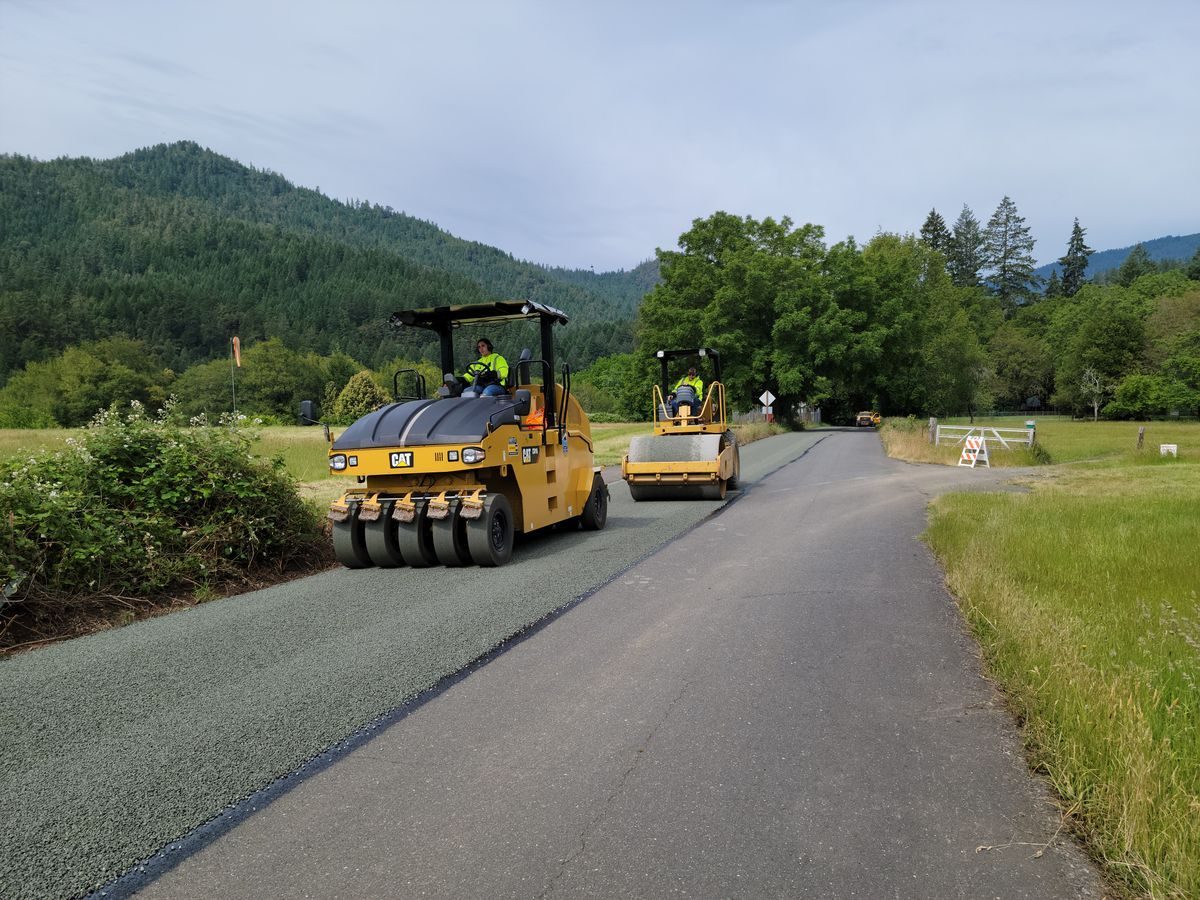 Road rollers on a newly treated lane beside green hills.