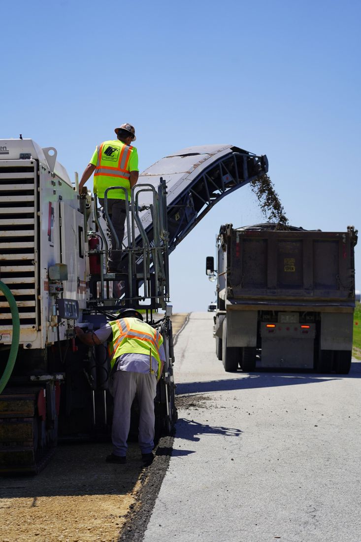 Workers operating a pavement milling machine that loads removed material into a truck.