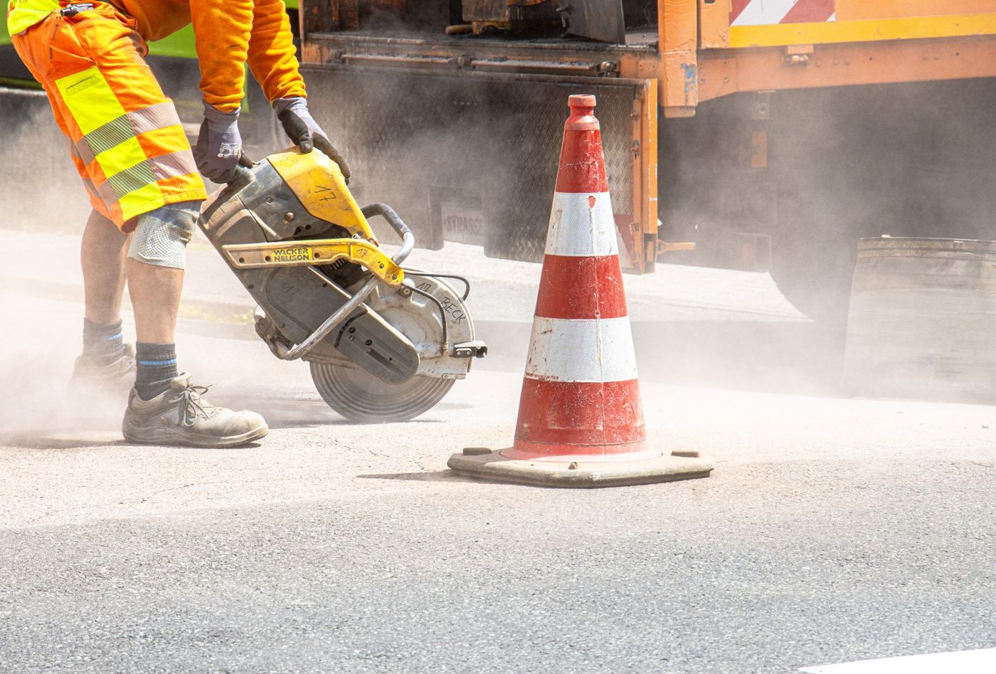 A worker saw-cutting pavement beside a traffic cone.