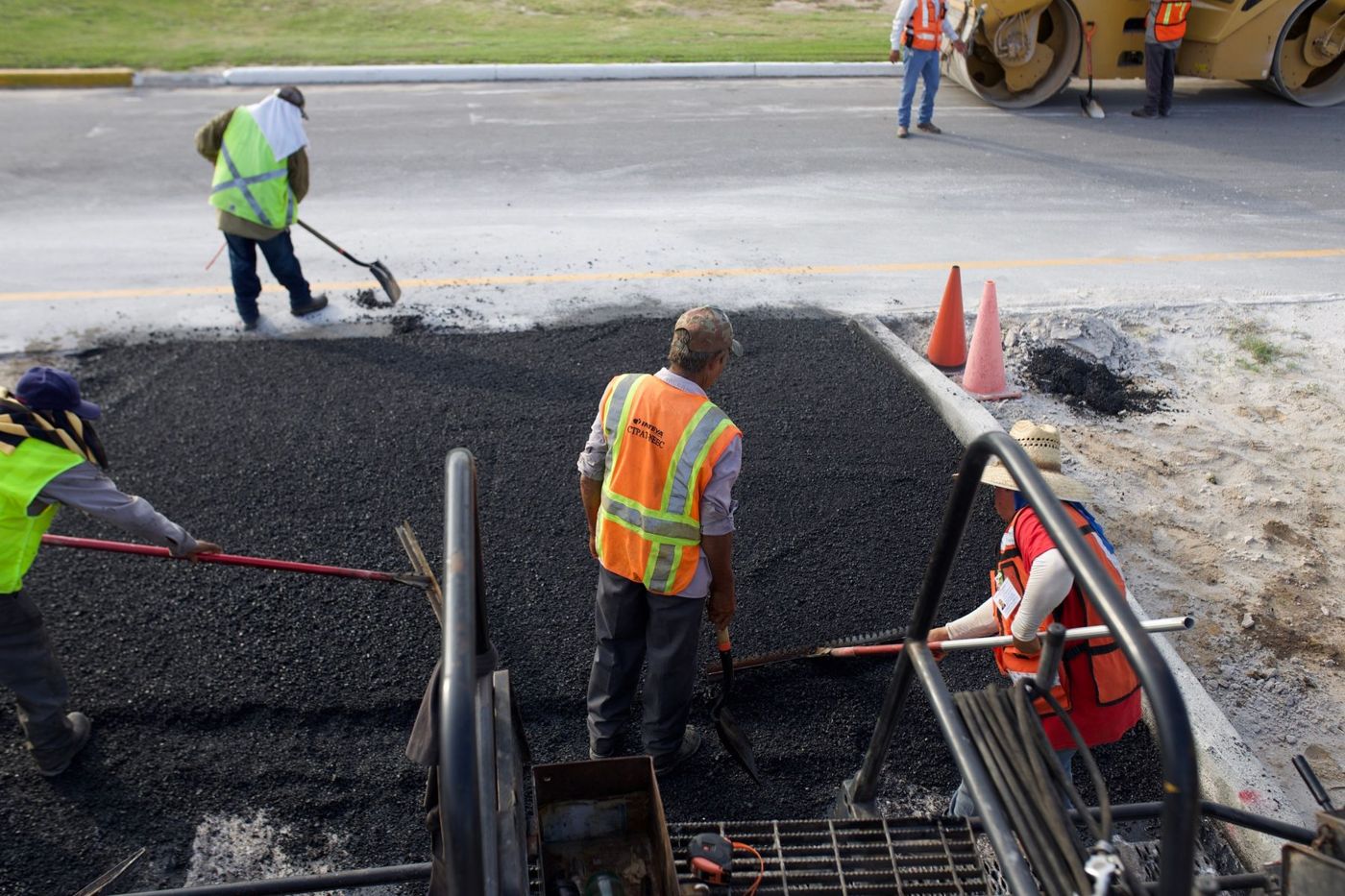 Road workers beside asphalt paving equipment and traffic cones.